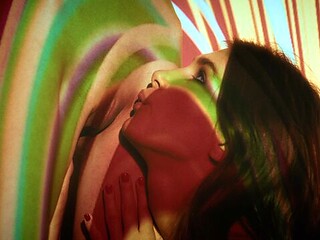 Ellie Luna and Leona Mia kiss slowly under swirling red and yellow light projections